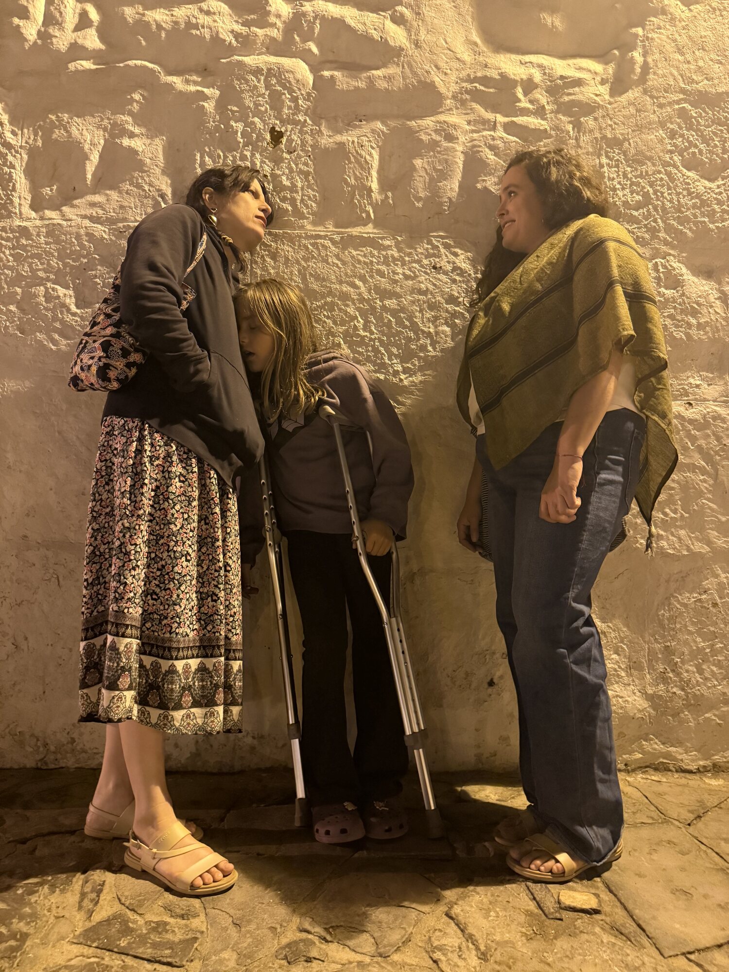 Figures against a weathered plaster wall in golden light — a woman and children, one holding crutches