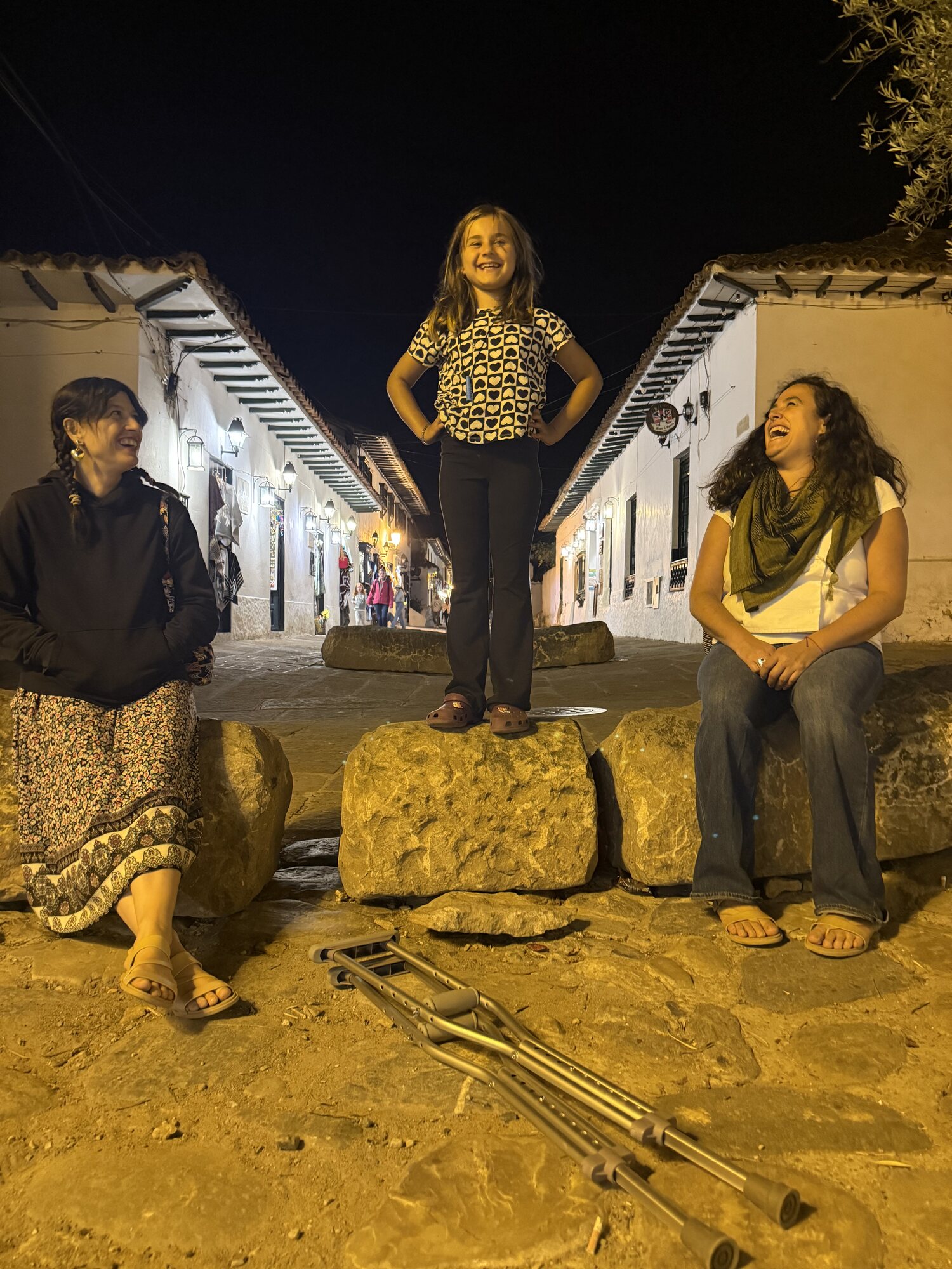 A girl stands triumphant on a stone at night in a colonial street, women around her, crutches resting on the ground