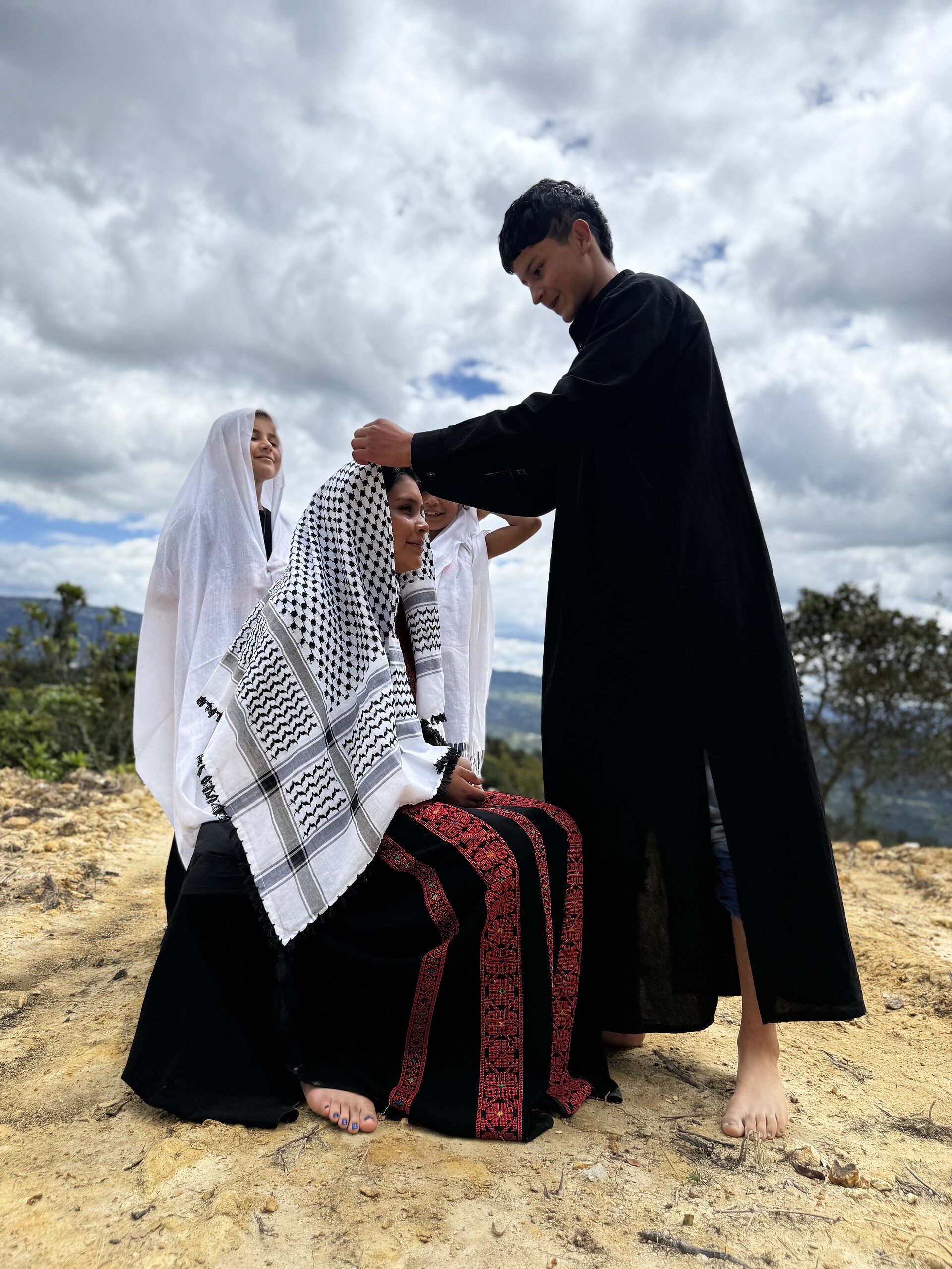 A boy in black drapes a keffiyeh over a seated woman while children in white veils look on, under storm clouds