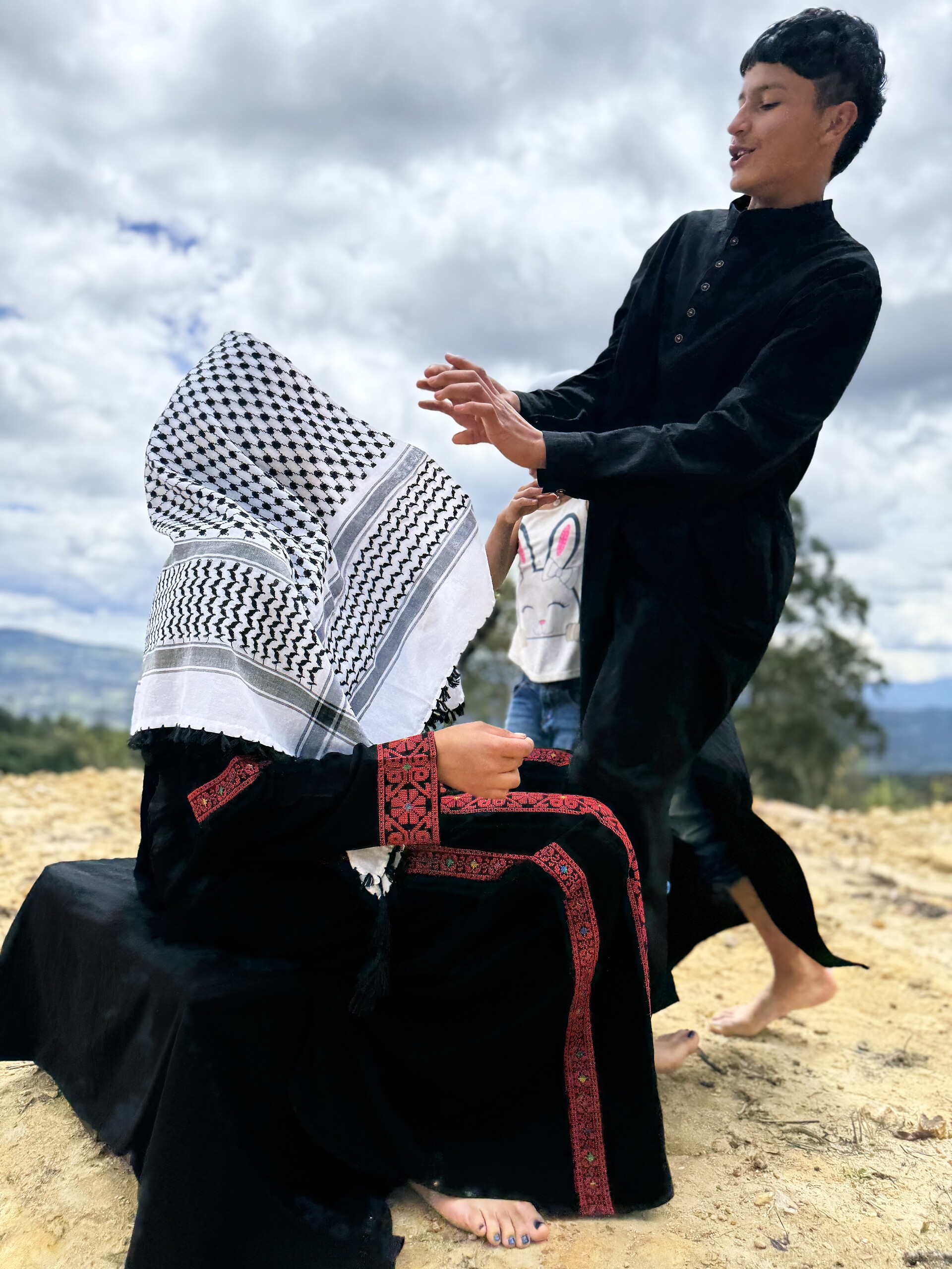 A boy in black moves with outstretched hands beside a seated figure veiled in a keffiyeh, under a storm sky