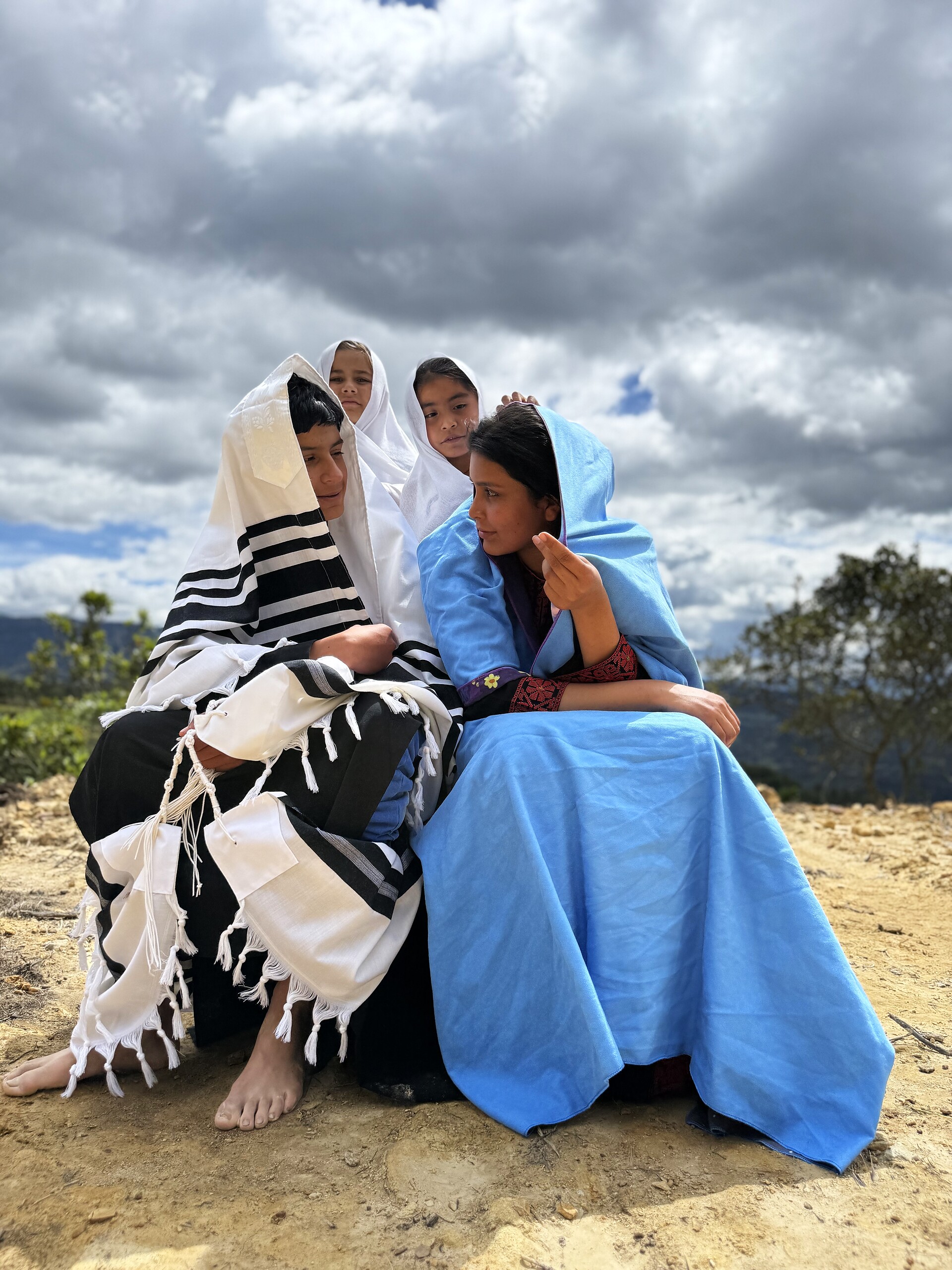 A group seated close together under storm clouds — a boy in a tallit, a girl in a blue veil, and children in white