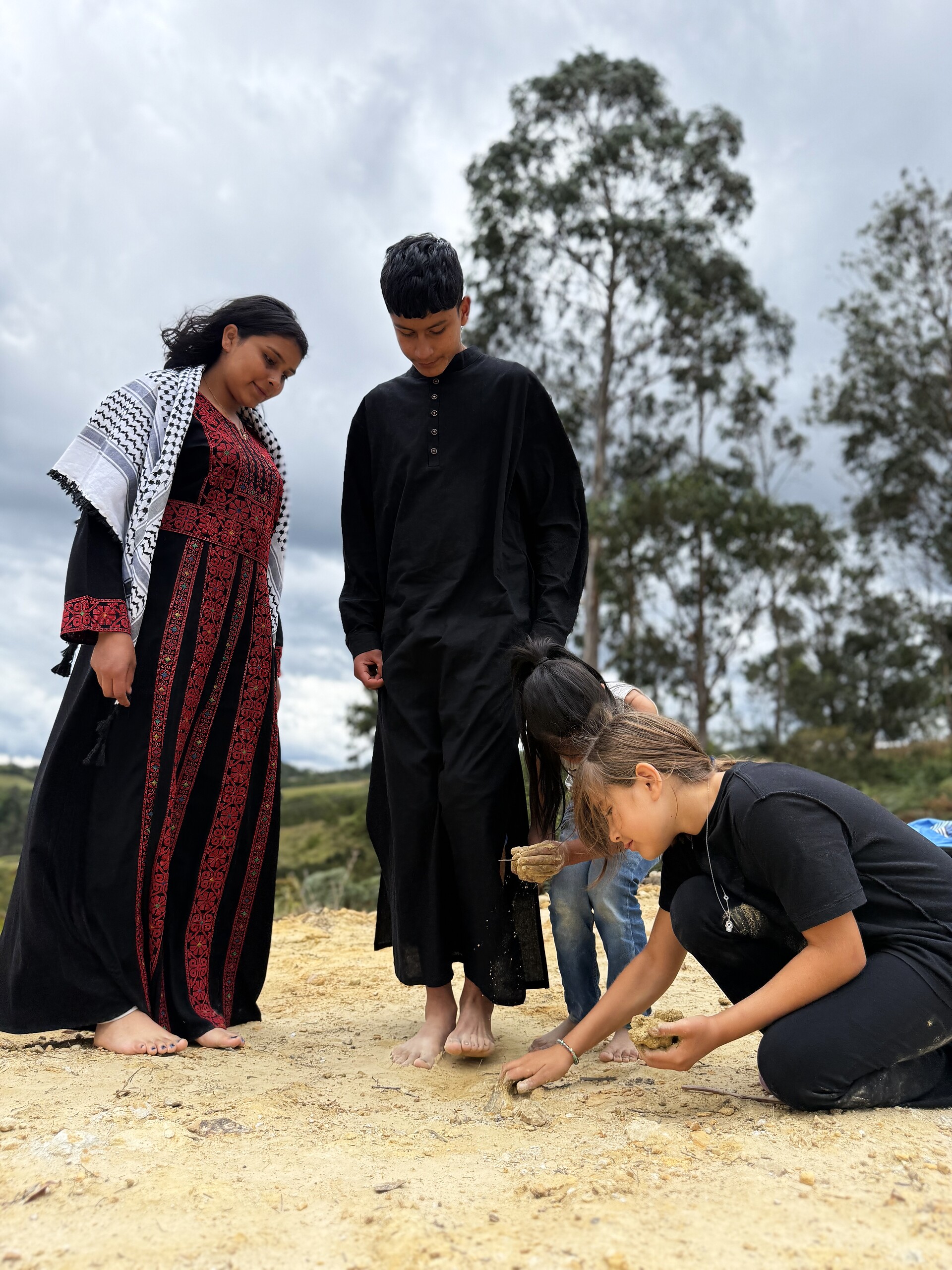 Two figures watch two children dig in the earth on a hilltop, under a grey sky