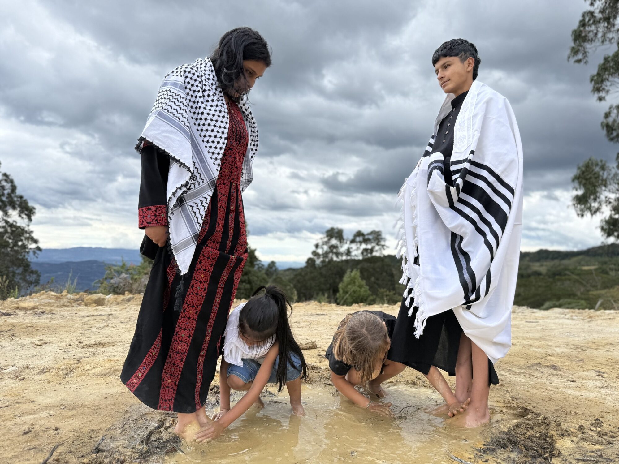 Two figures stand on a hilltop under a storm sky — one wrapped in a Palestinian keffiyeh and embroidered thobe, one in a Jewish tallit — while two children wash their hands together in a muddy pool between them.